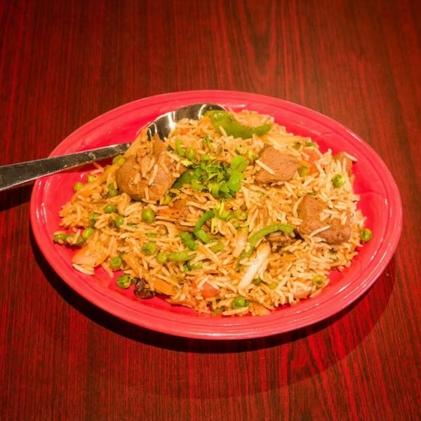 A vibrant red plate filled with biryani, featuring rice, peas, meat, and green garnishes, with a spoon.