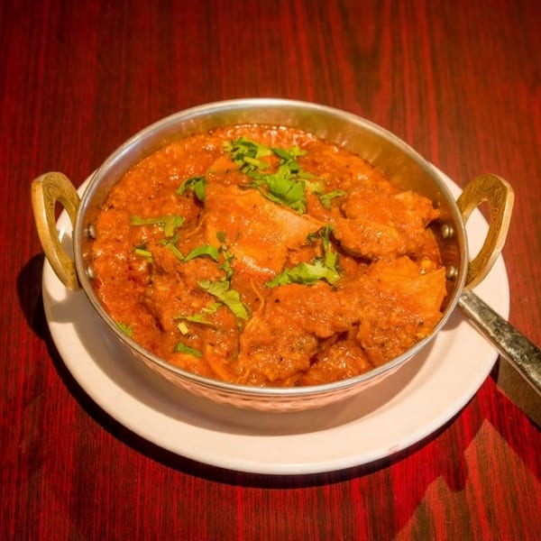 A metal bowl of chicken tikka masala, garnished with cilantro, on a white plate and red table.
