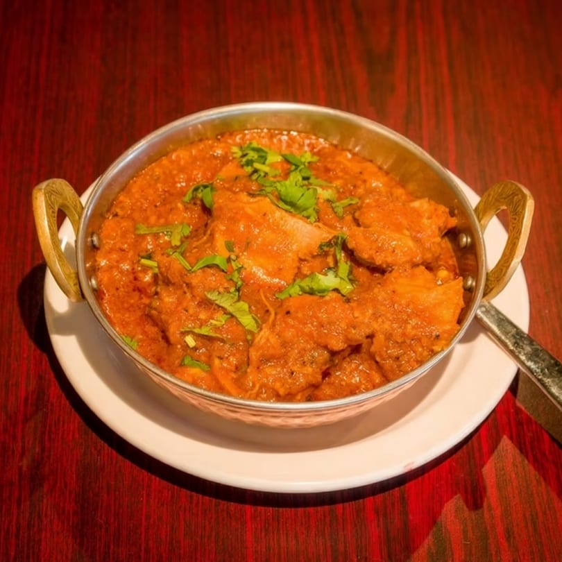 A metal bowl of chicken tikka masala, garnished with cilantro, on a white plate and red table.