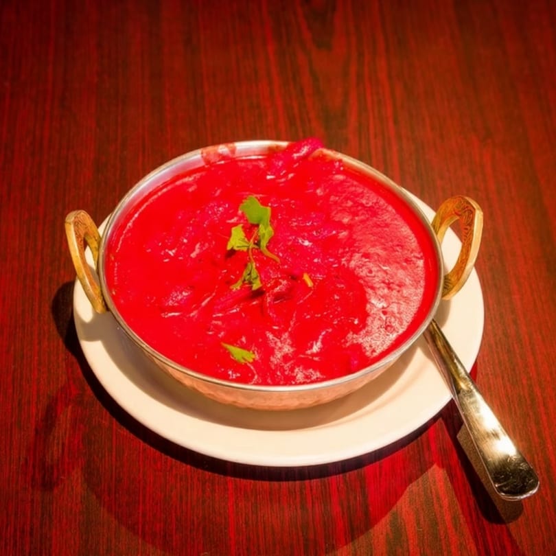 Vibrant red curry with green herbs in a brass bowl on a white plate and dark wooden table.