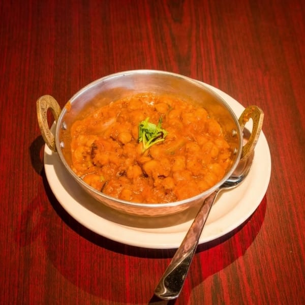 A metal bowl of chana masala, garnished with cilantro, sits on a white plate with a spoon.