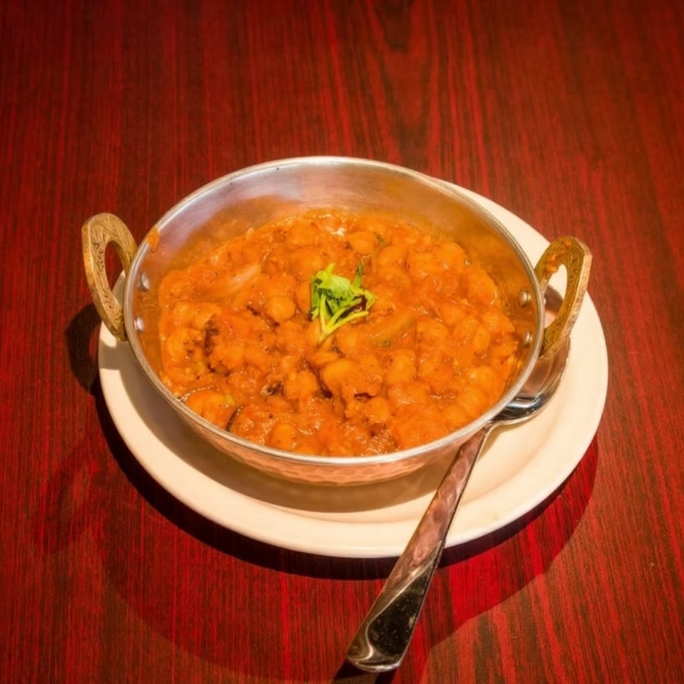 A metal bowl of chana masala, garnished with cilantro, sits on a white plate with a spoon.