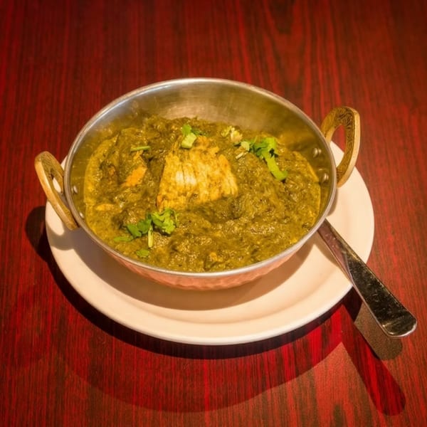 A metal bowl of vibrant green palak paneer, garnished with cilantro, sits on a white plate.