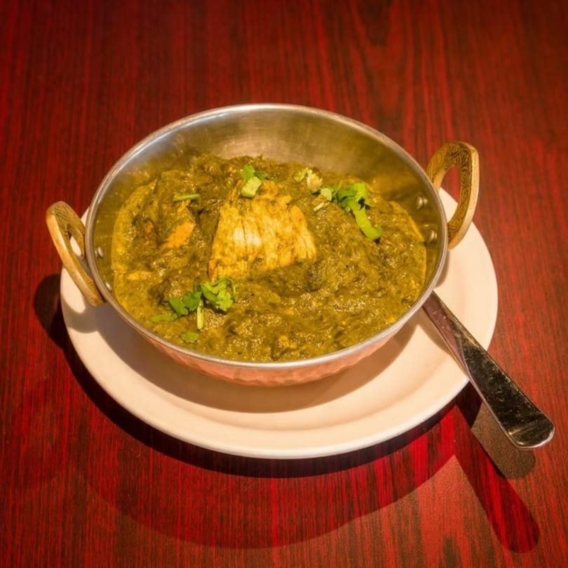 A metal bowl of vibrant green palak paneer, garnished with cilantro, sits on a white plate.