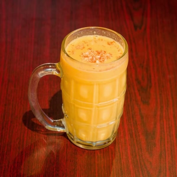 A glass mug of mango lassi, topped with nuts and spices, sits on a dark red wooden table.