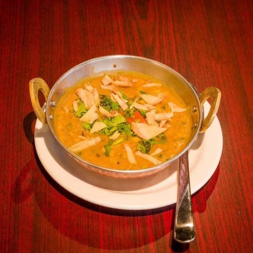 A metal bowl of yellow lentil soup with cilantro and flatbread pieces on a red wooden table.