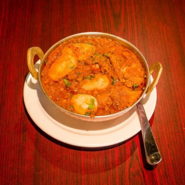 A metal bowl of Indian curry with potatoes and cilantro, on a white plate with a spoon.