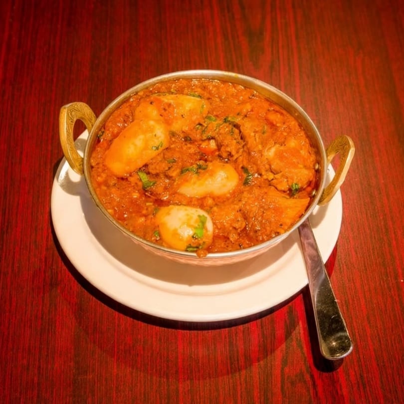 A metal bowl of Indian curry with potatoes and cilantro, on a white plate with a spoon.