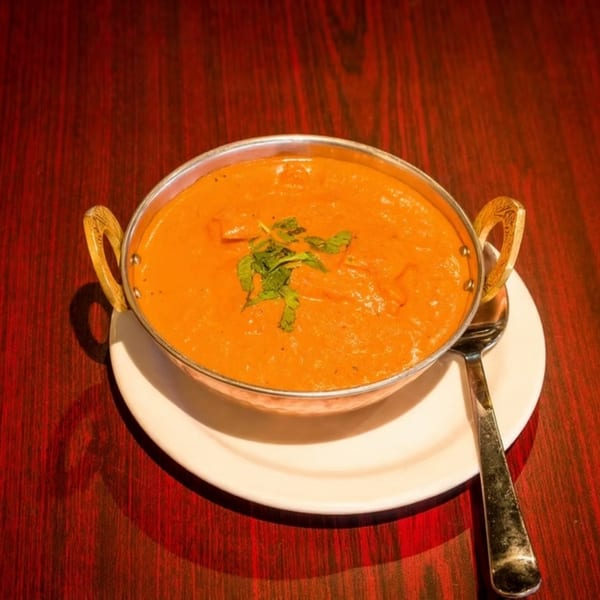 A copper bowl of orange curry with green garnish sits on a white plate and dark wooden table.